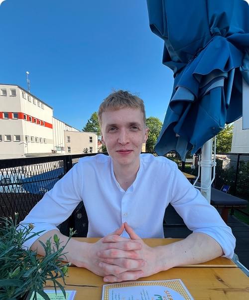 Młody blondyn w białej koszuli siedzi przy stoliku na tarasie, ręce splecione, uśmiechnięty, w tle niebieskie parasole i budynek, na stole menu i zielona roślinka.